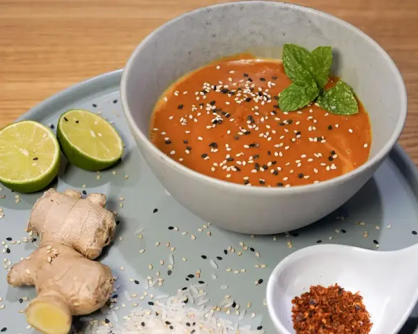 Schüssel mit cremiger Erdnusssauce, garniert mit Sesam und Minze. Daneben Limetten, Ingwer und Chiliflocken – Zutaten für ein leckeres Erdnusssauce Rezept.