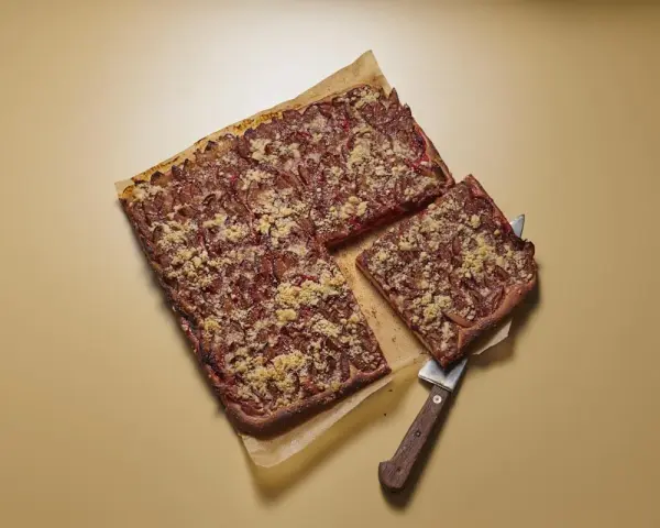 Ein rechteckiger Zwetschgenkuchen mit Streuseln, ein Stück wird mit einem Kuchenheber herausgehoben.