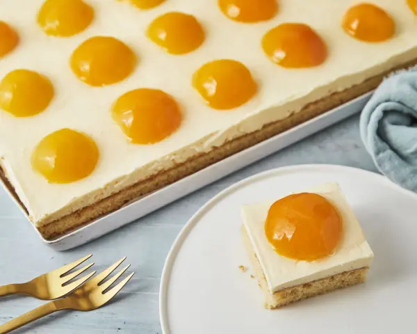 Ein rechteckiger Kuchen mit einer cremigen Schicht und vielen Pfirsichhälften, die wie ein Spiegelei Kuchen aussehen. Ein Stück auf einem Teller daneben.