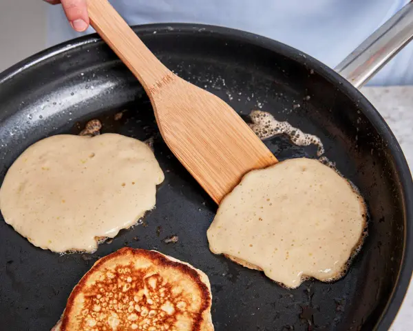 Buttermilch pancakes braten in der Pfanne. Ein Holzspatel wendet einen goldbraunen Pancake.