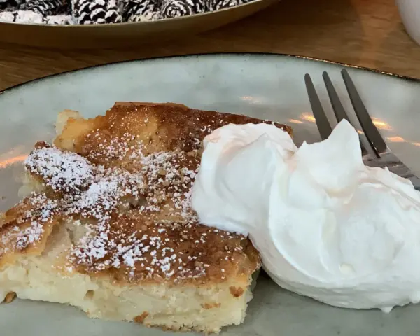 Ein Stück saftiger Apfelkuchen, bestäubt mit Puderzucker, serviert mit einer großen Portion Schlagsahne und einer Gabel.