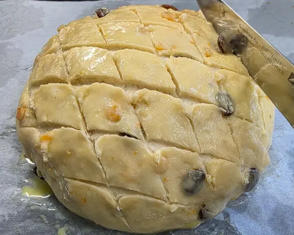 Ein runder Teigling mit eingeschnittenen Rauten, bereit für das Osterfladen Rezept. Rosinen und Orangenschale sind sichtbar.