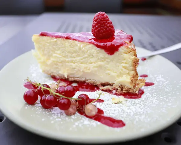 Ein Stück cremiger Käsekuchen mit Himbeersoße und frischen Beeren, bestäubt mit Puderzucker. Ein Genuss für alle Sinne, diese Himbeer Torte.
