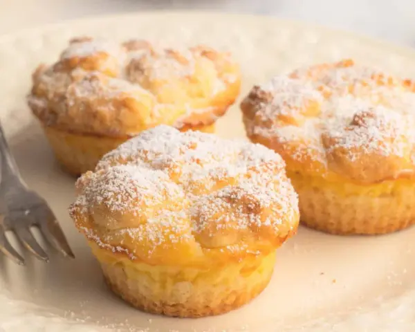 Drei luftige cheesecake muffins, bestäubt mit Puderzucker, warten auf einer Platte auf eine Gabel.