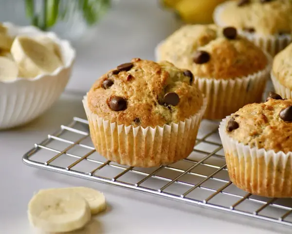 Frisch gebackene schoko bananen muffins mit Schokostückchen auf einem Gitter. Daneben eine Schüssel mit Bananenscheiben.