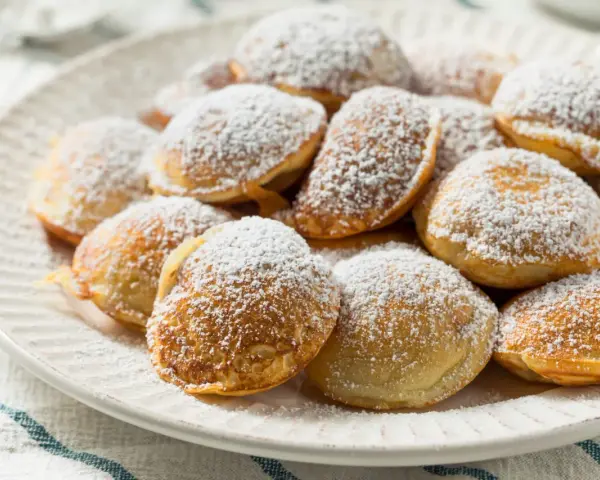 Ein Teller voller goldbrauner Poffertjes, bestäubt mit Puderzucker. Dieses Poffertjes Rezept ist ein Genuss!