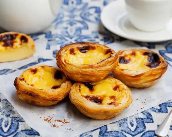 Vier köstliche Pastel de Nata auf einem blauen Tischtuch, mit einer Tasse Kaffee im Hintergrund.