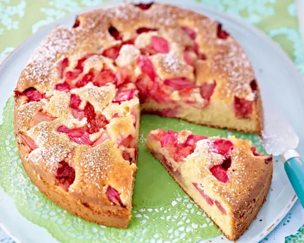 Ein saftiger Erdbeer Rhabarber Kuchen, bestäubt mit Puderzucker, liegt auf einem Teller. Ein Stück wurde bereits entnommen.