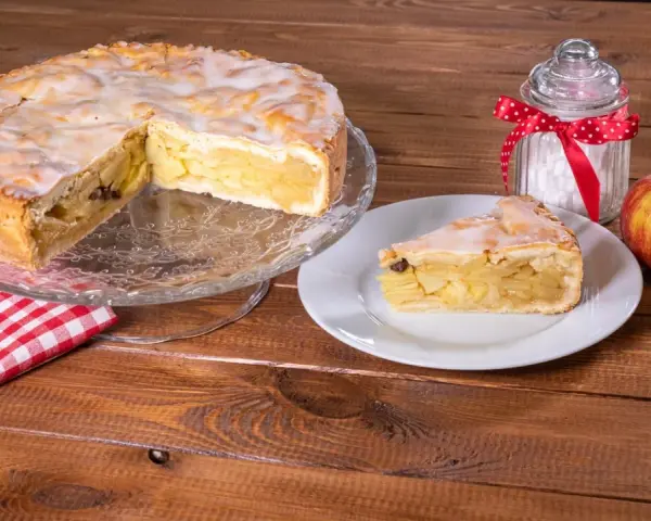 Omas weltbester Apfelkuchen, saftig und mit Zuckerguss überzogen, wartet auf einem Tortenständer. Ein Stück auf einem Teller und ein Apfel daneben laden zum Genießen ein.