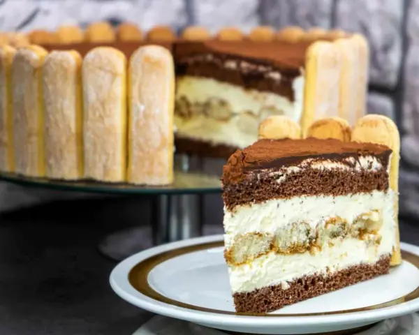 Ein Stück Tiramisu Torte mit Löffelbiskuits und Kakao bestäubt. Im Hintergrund die ganze Torte, ebenfalls mit Löffelbiskuits umrandet.