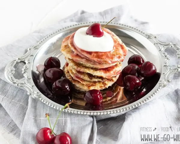 Ein Stapel Bananen Pancakes mit Kirschen und Sahne auf einem Silbertablett, garniert mit frischen Kirschen.