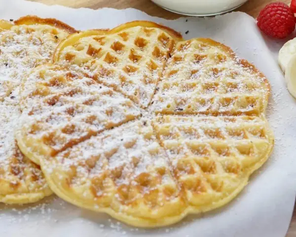 Zuckerbestäubte Buttermilchwaffeln, die auf einem Holzbrett liegen. Daneben sind Himbeeren und Bananenscheiben zu sehen.