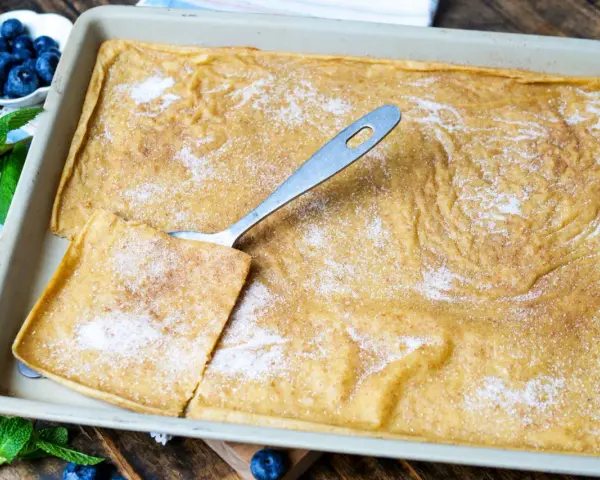 Ein frisch gebackener Ofenpfannkuchen, bestreut mit Zucker und Zimt, bereit zum Servieren. Ein Stück wird mit einem Löffel herausgehoben.