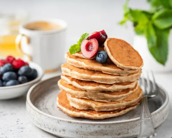 Ein Stapel veganer Pancakes, garniert mit Kirschen und Blaubeeren, daneben eine Schale Beeren und eine Tasse Kaffee.