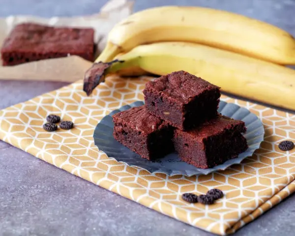 Saftige Bananen Brownies auf einem gelben Tuch, daneben reife Bananen. Ein Genuss!