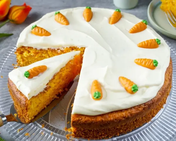Saftiger Karottenkuchen mit cremigem Frosting und süßen Marzipan-Karotten. Ein Stück ist bereits angeschnitten.