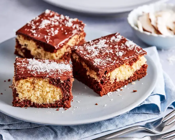 Drei Stücke saftiger Schoko Kokos Kuchen mit Schokoladenglasur und Kokosraspeln auf einem grauen Teller.