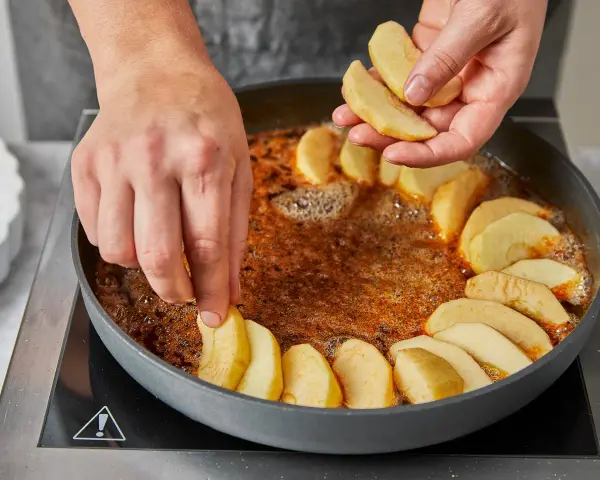 Hände legen Apfelspalten in eine Pfanne mit karamellisierter Butter. Die Zubereitung der klassischen Tarte Tatin beginnt.