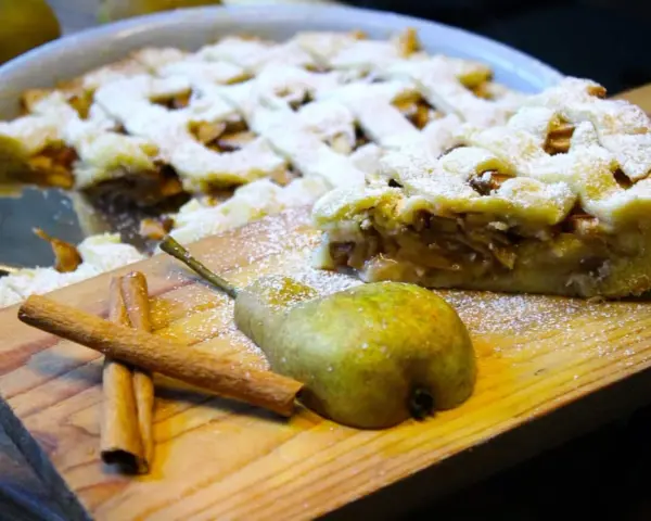 Ein köstlicher Birnenkuchen mit Gittermuster, bestäubt mit Puderzucker. Daneben liegen eine Birne und Zimtstangen – perfekt für ein Birnenkuchen Rezept.