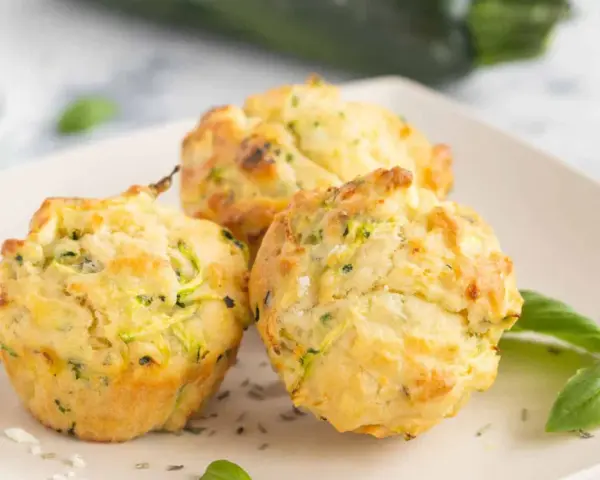 Drei goldbraune Zucchini-Muffins mit Kräutern und Käse auf einem Teller, garniert mit Basilikum.