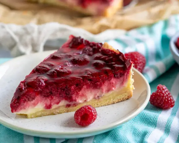 Ein saftiges Stück Himbeer Schmand Kuchen mit frischen Himbeeren und glänzender Glasur auf einem Teller.