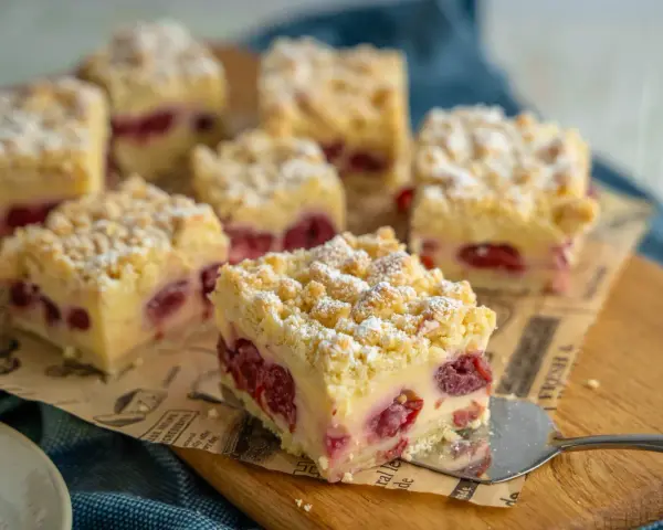 Saftiger Kirschkuchen mit Pudding und Streuseln, bestäubt mit Puderzucker. Ein Stück wird gerade serviert.