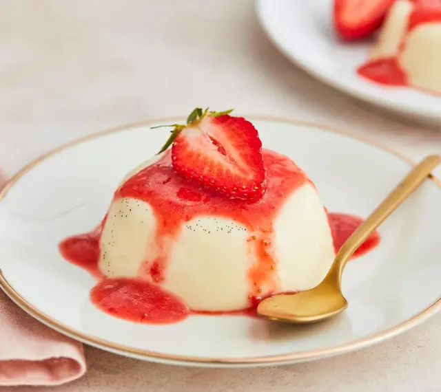 Cremige Panna Cotta mit Erdbeersauce und frischer Erdbeere, garniert mit Vanillepunkten. Eine goldene Löffel liegt daneben.