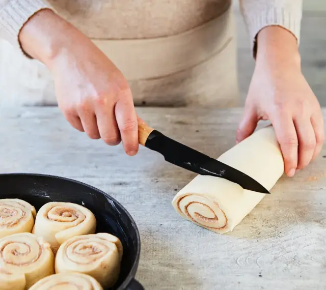 Hände schneiden Teigrolle für Zimtschnecken. Bereits geformte Schnecken warten in einer Pfanne auf das Backen. Ein tolles Zimtschnecken Rezept!