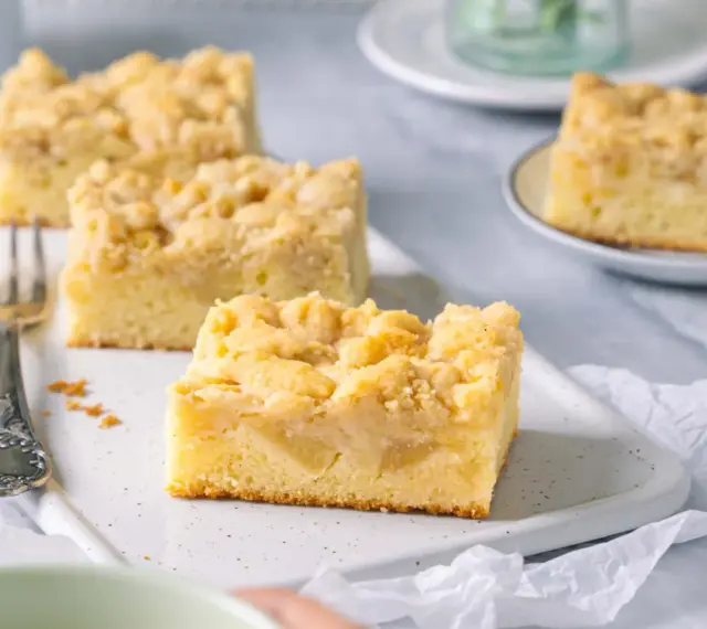 Saftiger Apfelkuchen mit Streuseln, perfekt für eine Kaffeepause. Ein Stück ist schon auf einer kleinen Platte angerichtet.