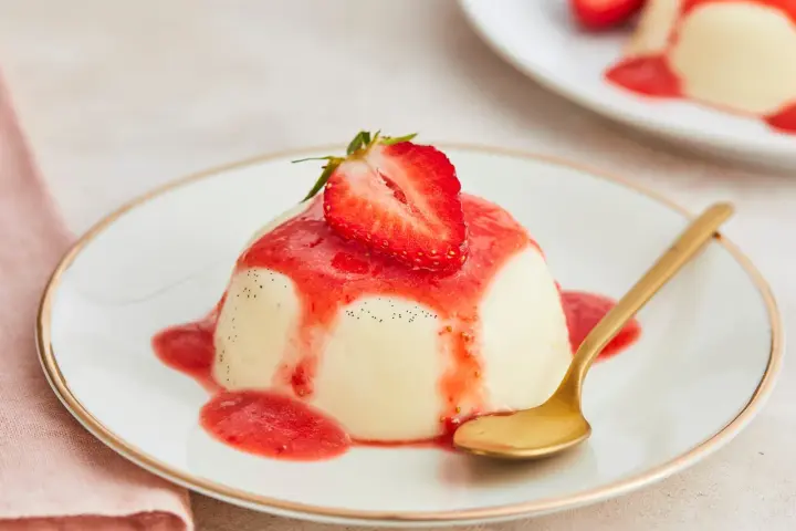 Cremige Panna Cotta mit Erdbeersauce und frischer Erdbeere, garniert mit Vanillepunkten. Eine goldene Löffel liegt daneben.