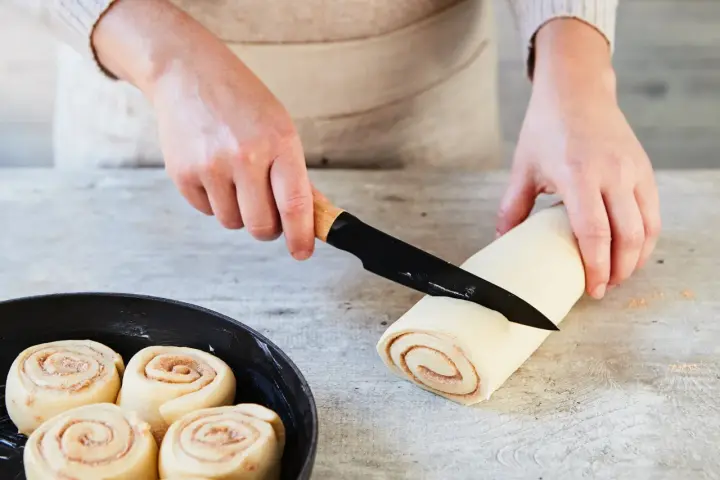 Hände schneiden Teigrolle für Zimtschnecken. Bereits geformte Schnecken warten in einer Pfanne auf das Backen. Ein tolles Zimtschnecken Rezept!
