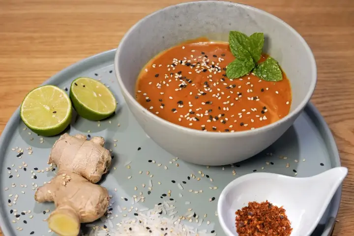 Schüssel mit cremiger Erdnusssauce, garniert mit Sesam und Minze. Daneben Limetten, Ingwer und Chiliflocken – Zutaten für ein leckeres Erdnusssauce Rezept.