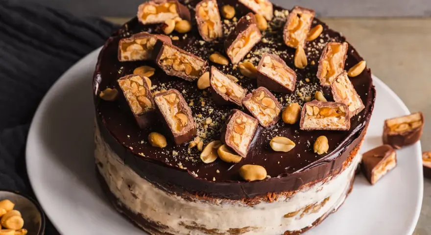 Eine köstliche Snickers Torte mit Schokoladenglasur, Erdnüssen und vielen Snickers-Stücken obendrauf.