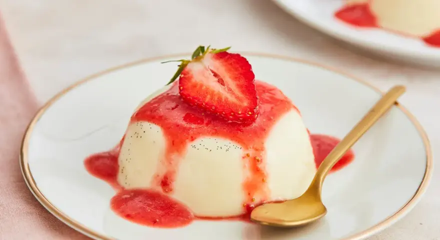 Cremige Panna Cotta mit Erdbeersauce und frischer Erdbeere, garniert mit Vanillepunkten. Eine goldene Löffel liegt daneben.