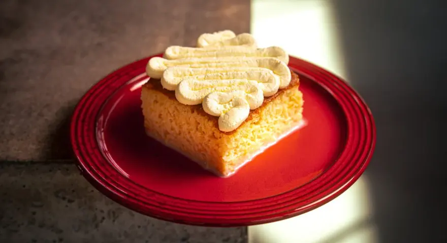 Ein Stück saftiger Tres Leches Kuchen mit Sahnetupfern auf einem roten Teller, bereit zum Genießen.
