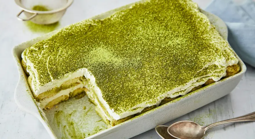 Ein cremiges Matcha Tiramisu, bestäubt mit feinem grünen Pulver, wartet in einer Auflaufform darauf, genossen zu werden. Löffel liegen bereit.