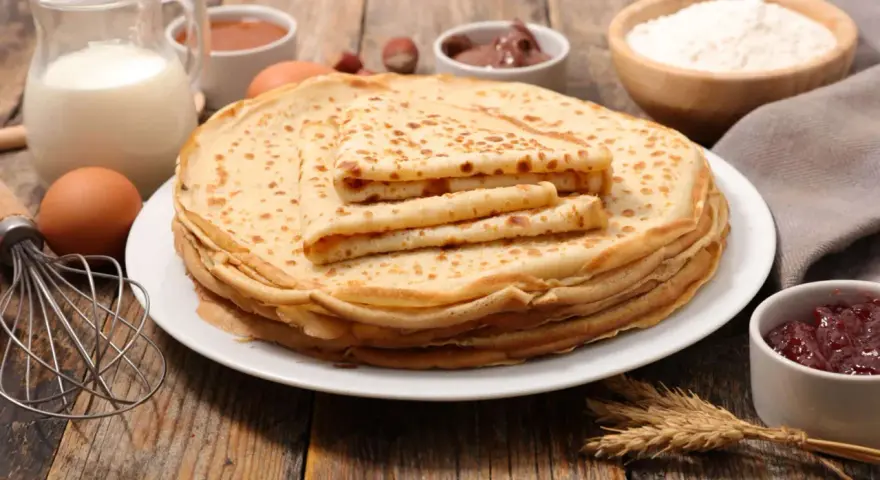 Ein Stapel goldbrauner Pfannkuchen, zubereitet nach einem sehr guten Pfannkuchen Rezept, daneben Zutaten wie Milch, Eier, Mehl, Marmelade und Nuss-Nougat-Creme.