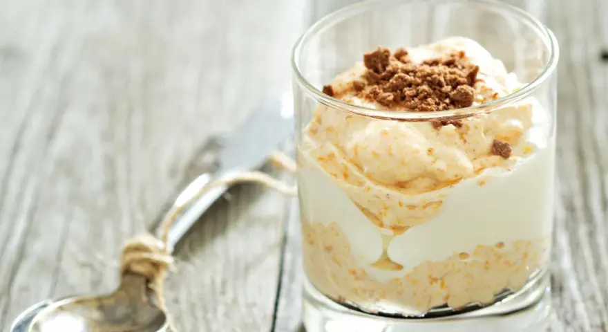 Cremiger cheesecake im glas, geschichtet mit weißer Creme und garniert mit zerbröselten Keksen. Ein Löffel liegt daneben.