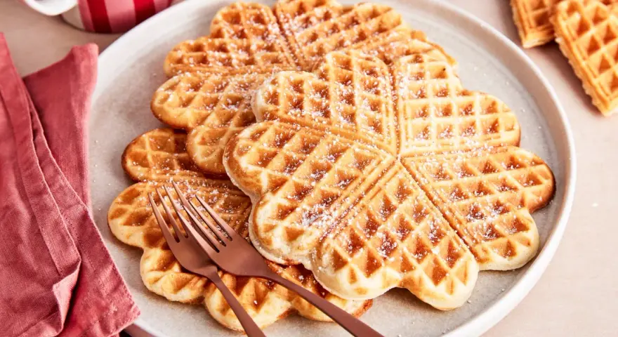 Herzförmige Waffeln, bestäubt mit Puderzucker. Ein köstlicher waffelteig ohne butter, serviert auf einem Teller mit zwei Gabeln.
