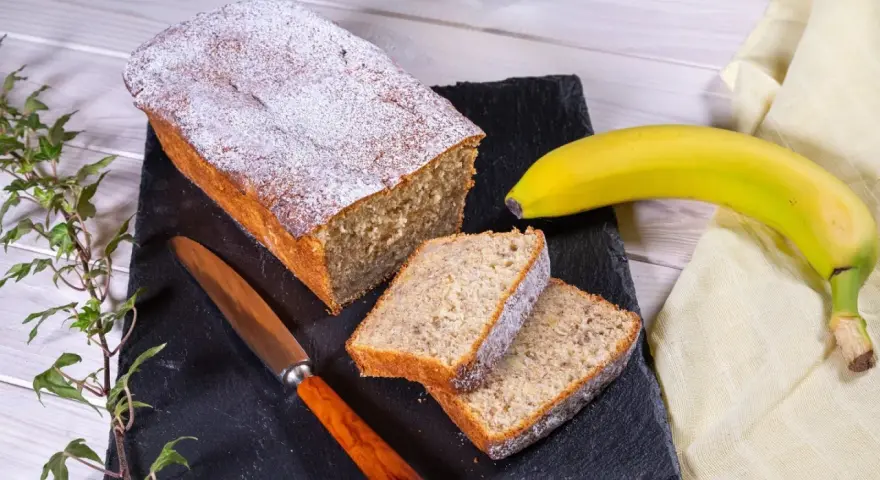 Saftiger Bananenkuchen, bestäubt mit Puderzucker, neben einer Banane und einem Messer. Perfekt für ein Bananenkuchen Rezept.