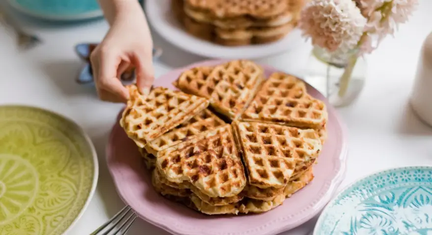 Ein Stapel herzförmiger Waffeln ohne Ei auf einem rosa Teller, bereit zum Genießen. Daneben liegen bunte Teller und eine Gabel.