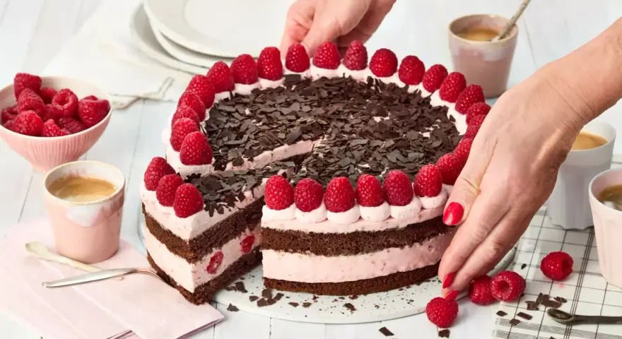 Eine köstliche Schoko Himbeer Torte, verziert mit frischen Himbeeren und Schokostreuseln, wird von Händen arrangiert.