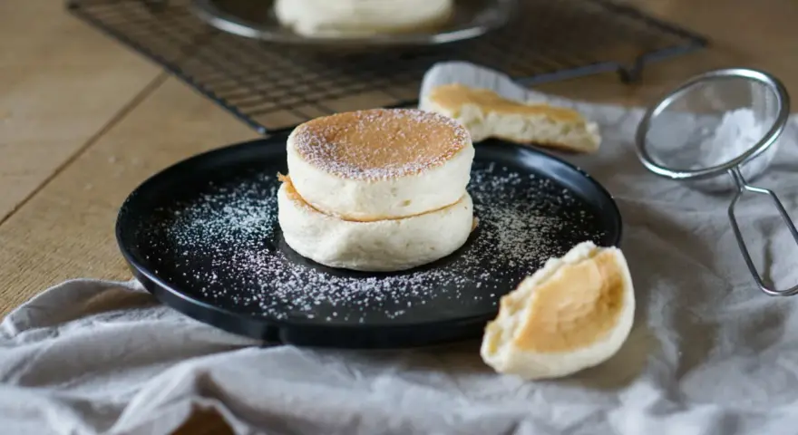 Zwei fluffige japanische Pancakes auf einem schwarzen Teller, bestäubt mit Puderzucker. Daneben ein Stück Pancake und ein Sieb.
