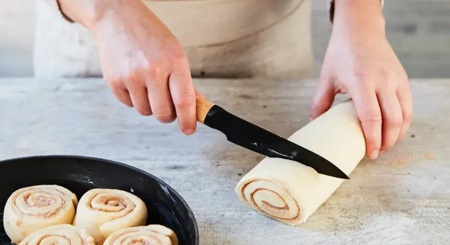 Hände schneiden Teigrolle für Zimtschnecken. Bereits geformte Schnecken warten in einer Pfanne auf das Backen. Ein tolles Zimtschnecken Rezept!