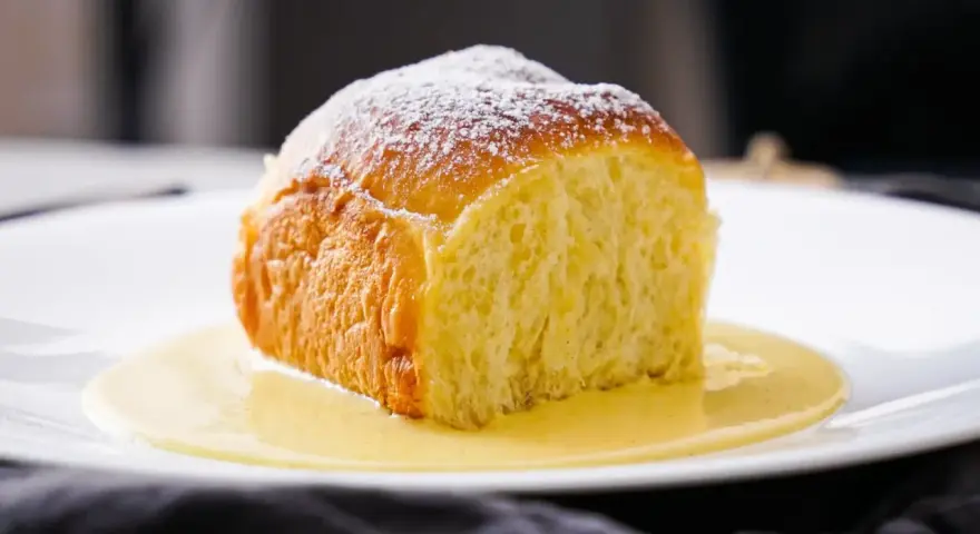 Ein goldbraunes Buchtel, mit Puderzucker bestäubt, ruht in einer cremigen Vanillesauce. Ein köstliches Buchtel Rezept zum Nachbacken.