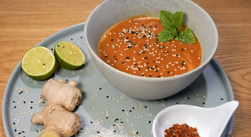 Schüssel mit cremiger Erdnusssauce, garniert mit Sesam und Minze. Daneben Limetten, Ingwer und Chiliflocken – Zutaten für ein leckeres Erdnusssauce Rezept.