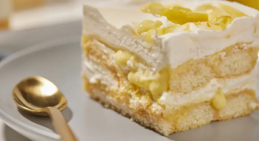 Ein Stück Zitronen Tiramisu mit cremiger Füllung und Zitronenscheiben, daneben eine goldene Löffel.