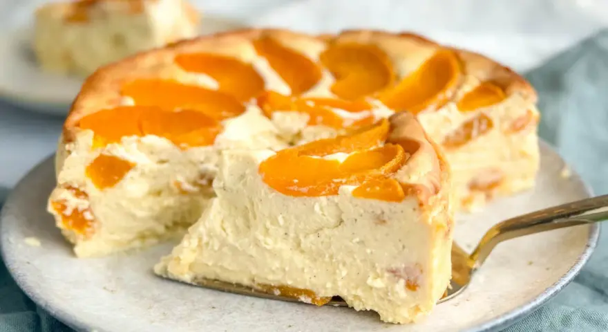Ein saftiger Pfirsichkuchen mit cremiger Füllung und frischen Pfirsichspalten. Ein Stück wird gerade mit einem goldenen Tortenheber herausgehoben.