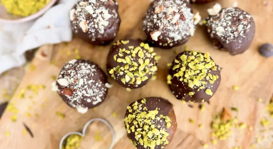 Selbstgemachte Pralinen mit Schokolade und gehackten Nüssen, einige mit Pistazien bestreut. Ein Herz-Ausstecher liegt daneben.