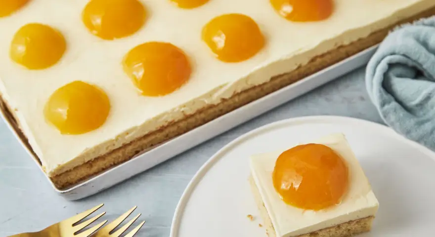 Ein rechteckiger Kuchen mit einer cremigen Schicht und vielen Pfirsichhälften, die wie ein Spiegelei Kuchen aussehen. Ein Stück auf einem Teller daneben.
